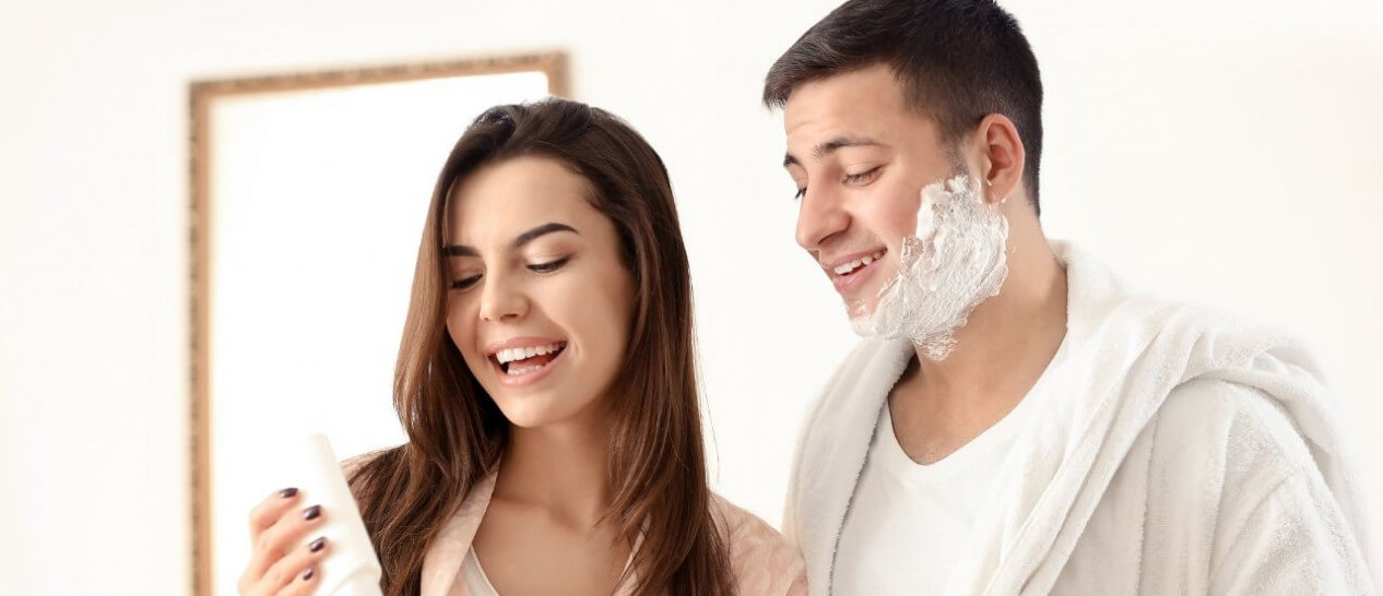 young couple laughing, man is shaving, woman is using losion